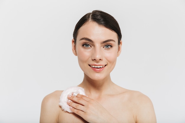 Beauty portrait of an attractive young brunette woman standing isolated over white wall, touching body with powder puff