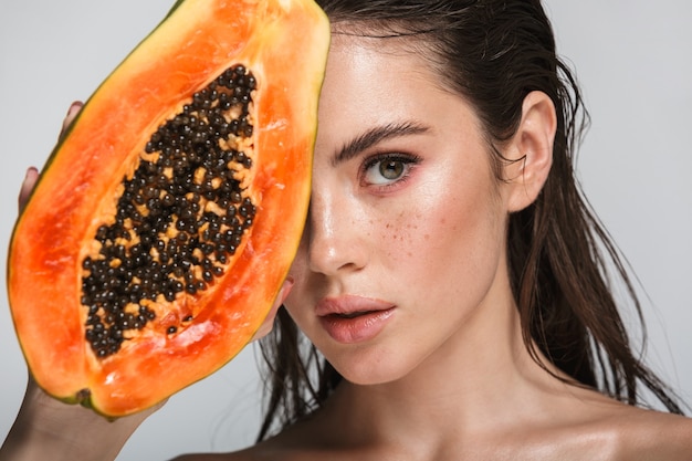 Beauty portrait of an attractive young topless brunette woman standing isolated on white, posing with papaya