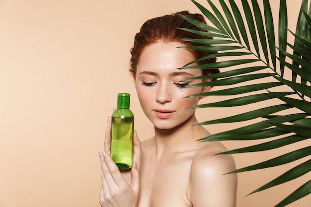 Beauty portrait of an attractive young topless redhead woman standing isolated, showing cosmetic lotion bottle, posing surrounded with tropical leaves