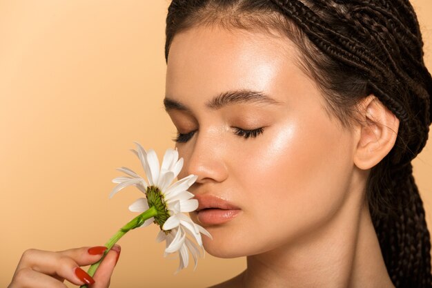 Beauty portrait of an attractive young topless woman standing isolated over beige wall, smelling camomile flower