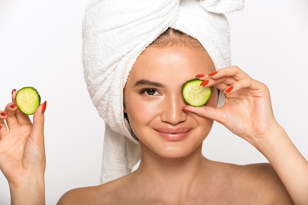 Beauty portrait of an attractive young topless woman wearing bathroom towel on her head standing isolated over white wall, holding cucumber slices at her eyes