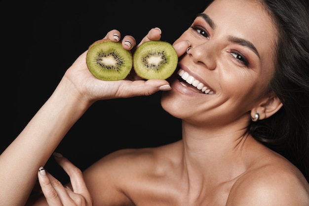 Beauty portrait of an attractive young topless woman with long brunette hair isolated over black wall, posing with sliced kiwi fruit