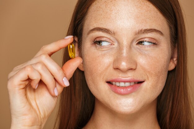 Ritratto di bellezza di una bella giovane donna in buona salute con lunghi capelli rossi che mostra la capsula vitaminica isolata sopra la parete beige