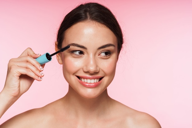 Beauty portrait of a beautiful smiling young topless brunette woman standing isolated on pink, applying mascara