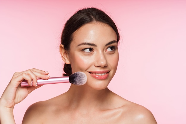 Beauty portrait of a beautiful smiling young topless brunette woman standing isolated on pink, holding makeup brush at her face