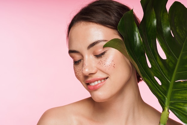 Beauty portrait of a beautiful smiling young topless brunette woman standing isolated on pink, posing with green tropical leaf