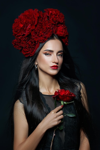 Beauty portrait brunette woman with crown of roses flowers on\
her head. bright red makeup and lipstick. rose flowers in the hands\
of a woman