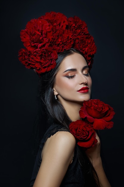 Beauty portrait brunette woman with crown of roses flowers on\
her head. bright red makeup and lipstick. rose flowers in the hands\
of a woman