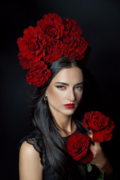 Женщина брюнет портрета красоты с короной цветков роз на ее голове. Ярко-красный макияж и помада. Розовые цветы в руках женщины