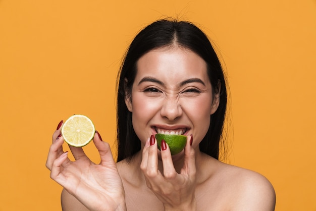 Beauty portrait of caucasian young brunette half-naked woman smiling and eating lemon isolated over yellow wall