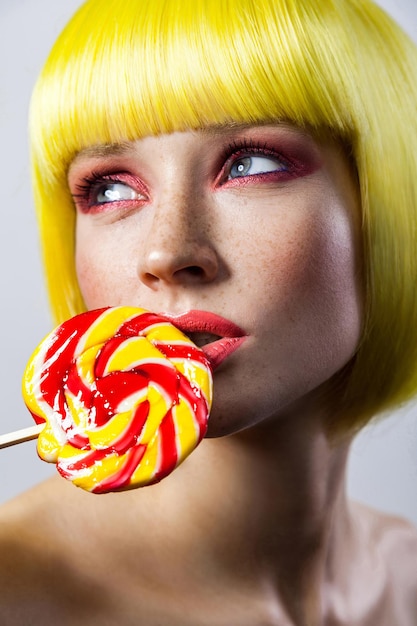 Beauty portrait of cute young fashion model with freckles, red makeup, yellow wig, holding colorful candy stick on lips and looking away with passion . indoor studio shot, isolated on gray background.