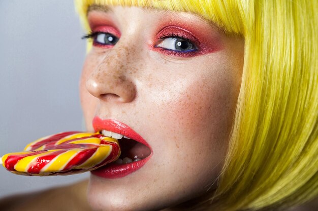 Beauty portrait of cute young female model with freckles, red makeup and yellow wig, holding and bitting her colorful candy stick and looking at camera. indoor studio shot, isolated on gray background