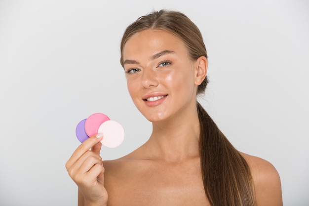 Beauty portrait of european shirtless woman with long brown hair smiling and holding colorful cosmetic pads, isolated