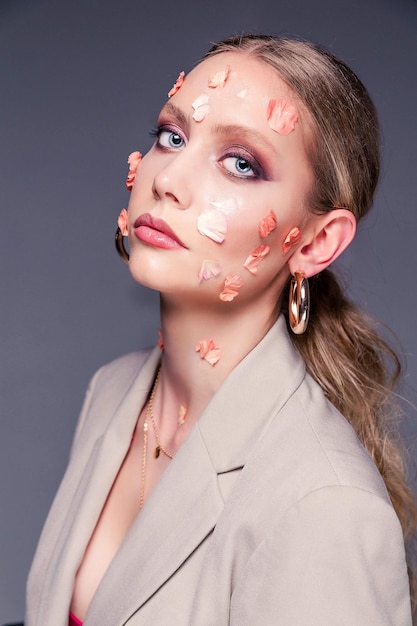 Beauty portrait of a girl Beautiful woman in a jacket posing with flower petals on her face on a gray background Femininity and beauty