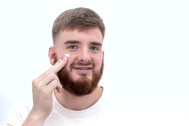 Beauty portrait of handsome happy young bearded man with beard and perfect skin on white background holding in hand cosmetic product applying cream on face smiling Male skin care Moisturizing