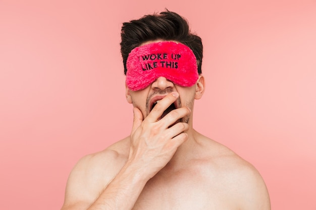 Beauty portrait of a handsome tired shirtless brunette man standing isolated over pink wall, wearing sleeping mask, yawning