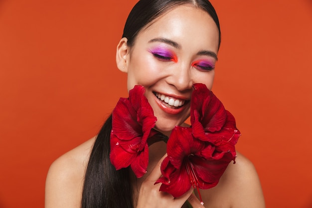 Beauty portrait of a happy young topless asian woman with brunette hair wearing bright makeup, standing isolated on red, posing with a red flower