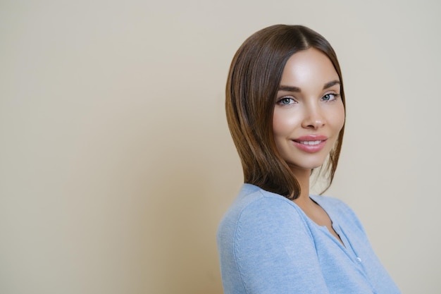 Beauty portrait of healthy lovely gorgeous woman with gentle smile clean healthy skin stands sideways has pleasant talk with someone looks directly at camera isolated over beige wall empty space