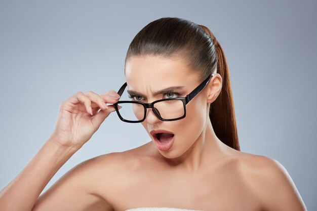 Beauty portrait of irritated girl in glasses looking aside aggressively. Head and shoulders of angry woman with opened mouth, touching glasses, staring