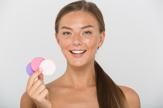 Photo beauty portrait of lovely shirtless woman with long brown hair smiling and holding colorful cosmetic pads, isolated