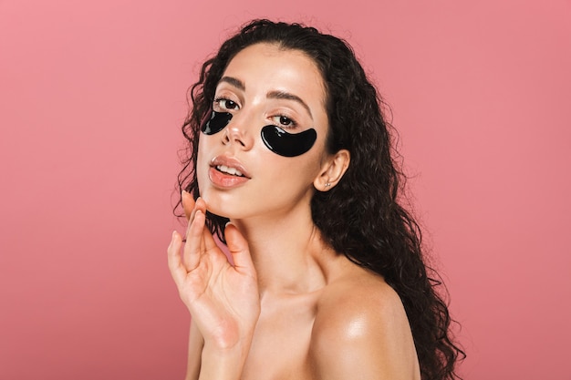 Beauty portrait of lovely shirtless woman with long brunette hair wearing cosmetic patches under her eyes, isolated over pink wall