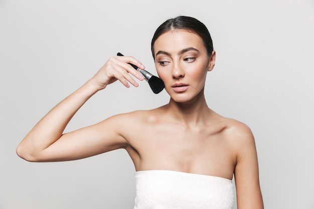 Портрет красоты молодой здоровой привлекательной брюнетки женщины, стоящей изолированно, применяя макияж с помощью кисти