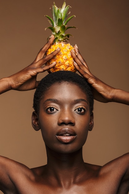 Beauty portrait of pretty young half-naked african woman holding pineapple on her head isolated on brown