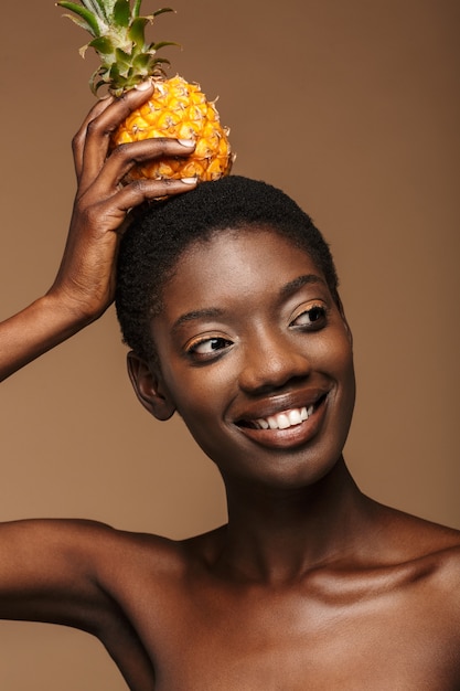 Beauty portrait of pretty young half-naked african woman holding pineapple on her head isolated on brown