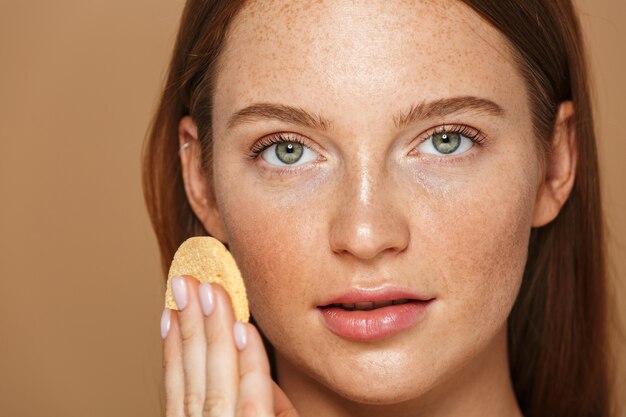 Beauty portrait of a smiling young topless woman with long red hair cleansing her face with a sponge isolated over beige wall