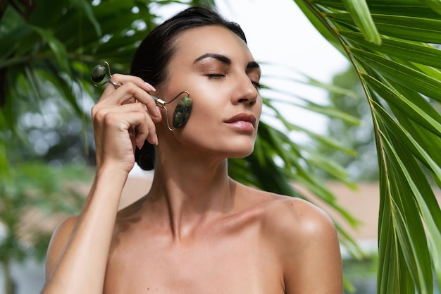 Beauty portrait soft skin Female model with natural makeup and healthy skin behind a green leafy plant Portrait of a beautiful girl with nude nails bare shoulders holding roller massager outdoor