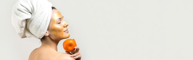 Beauty portrait of woman in white towel on head with gold nourishing mask on face Skincare cleansing eco organic cosmetic spa relax concept A girl stands with her back holding an orange mandarin