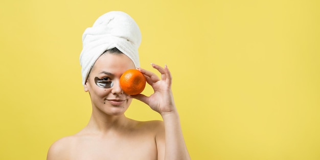 顔にゴールドの栄養マスクを頭に白いタオルで女性の美しさの肖像画スキンケアクレンジングエコオーガニック化粧品スパリラックスコンセプト女の子はオレンジ色のマンダリンを持って背中を持って立っています