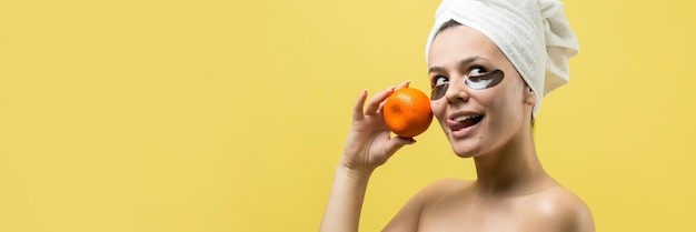 Beauty portrait of woman in white towel on head with gold nourishing mask on face Skincare cleansing eco organic cosmetic spa relax concept A girl stands with her back holding an orange mandarin