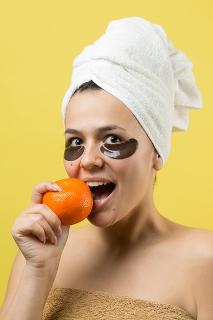 Beauty portrait of woman in white towel on head with gold nourishing mask on face. Skincare cleansing eco organic cosmetic spa relax concept. A girl stands with her back holding an orange mandarin.