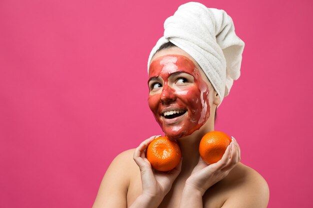 Beauty portrait of woman in white towel on head with red nourishing mask on face Skincare cleansing eco organic cosmetic spa relax concept A girl stands with her back holding an orange mandarin