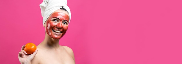 Beauty portrait of woman in white towel on head with red nourishing mask on face Skincare cleansing eco organic cosmetic spa relax concept A girl stands with her back holding an orange mandarin