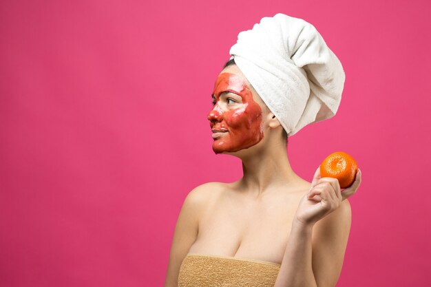 Beauty portrait of woman in white towel on head with red nourishing mask on face. Skincare cleansing eco organic cosmetic spa relax concept. A girl stands with her back holding an orange mandarin.