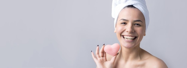 Beauty portrait of woman in white towel on head with a sponge for a body in view of a pink heart Skincare cleansing eco organic cosmetic spa relax concept