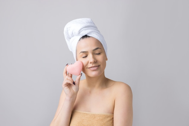 Beauty portrait of woman in white towel on head  with a sponge for a body in view of a pink heart. Skincare cleansing eco organic cosmetic spa relax concept.