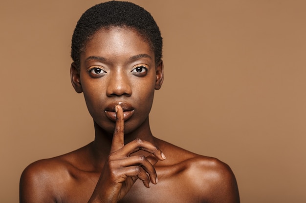 Beauty portrait of young half-naked african woman holding finger on lips isolated on beige