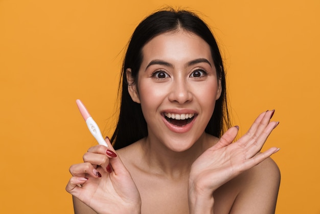 Beauty portrait of young half-naked woman rejoicing while holding pregnancy test isolated over yellow wall