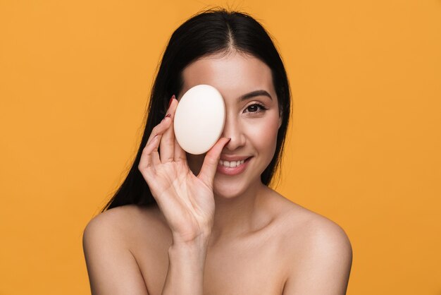 Beauty portrait of young half-naked woman smiling and holding bath soap isolated over yellow wall