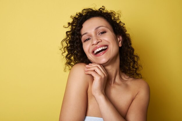 Beauty portrait of young half naked woman with curly hair smiling toothy smile looking at camera, posing over yellow all with copy space