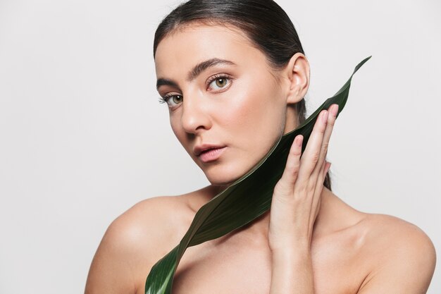 Beauty portrait of a young healthy attractive brunette woman standing isolated, posing with green tropical leaf