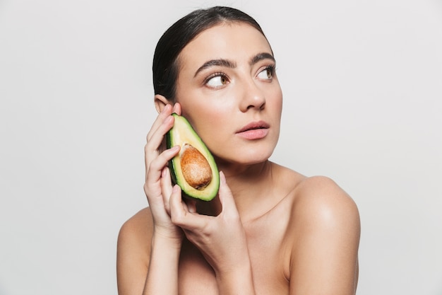 Beauty portrait of a young healthy attractive brunette woman standing isolated, posing with a sliced avocado