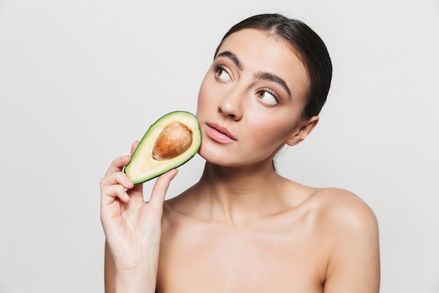 Портрет красоты молодой здоровой привлекательной брюнетки женщины, стоящей изолированно, позируя с нарезанным авокадо