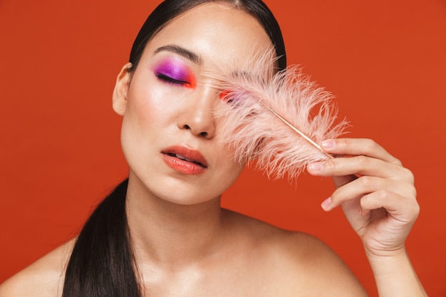 Beauty portrait of a young topless asian woman with brunette hair wearing bright makeup, standing isolated on red, holding a feather