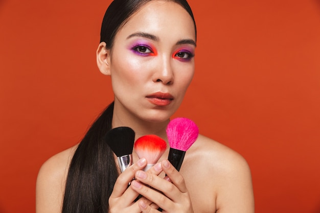 Портрет красоты молодой топлес азиатской женщины с волосами брюнетки, носящими яркий макияж, стоя изолированным на красном