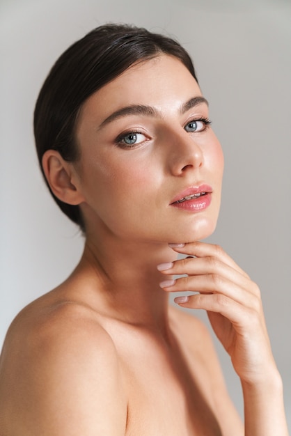 Beauty portrait of an young topless brunette woman standing isolated on gray, touching her face, looking at camera