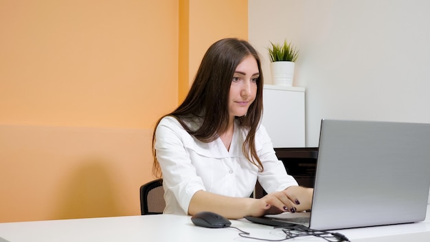 Beauty salon receptionist with long loose hair types on grey\
modern laptop against pot plant